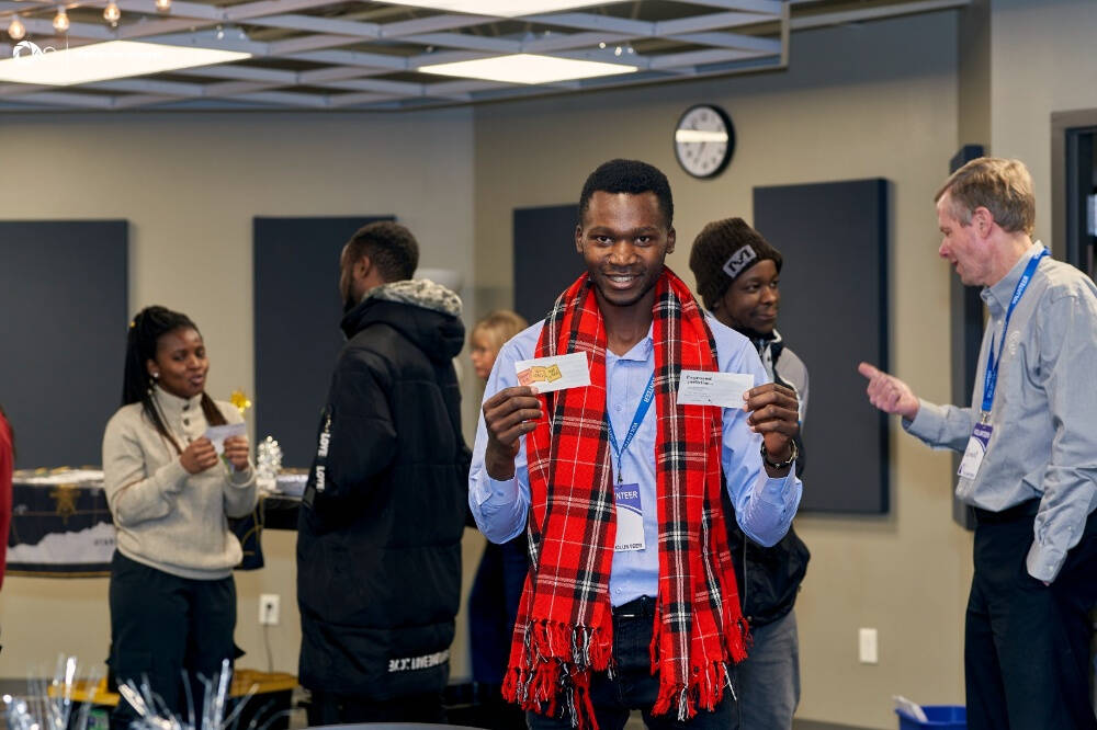 Erick Anangwe is wearing a blue shirt and a red Shuka- made in Kenya Scarf, and holds two cards, standing in a room with other people, some holding cards, against a backdrop of acoustic panels and a clock on the wall."